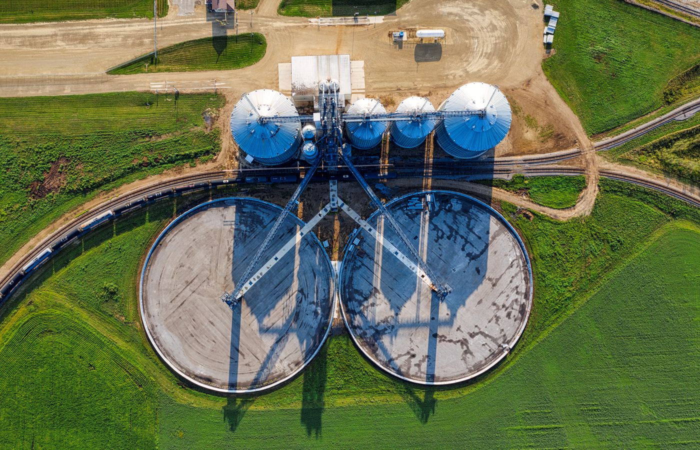 Grain silos and two round storage bunkers beside a railway, surrounded by green fields, seen from above.